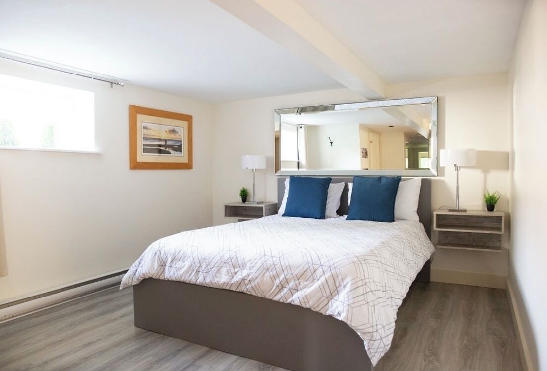A modern hotel room with a large bed, a bedside table with a lamp, a nightstand with a lamp, a framed picture on the wall, and a large mirror above the bed.