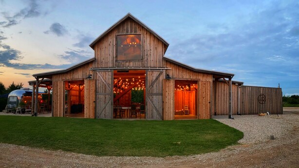 A rustic barn-style building with a large entrance, open interior, and a covered outdoor seating area. - The Range Vintage Trailer Resort (Ennis)