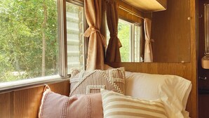 A cozy bedroom with a bed, pillows, a window with curtains, and a wall-mounted lamp. - The Range Vintage Trailer Resort (Ennis)