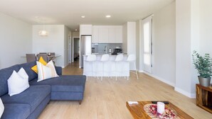 A modern living room with a grey sectional sofa, a small wooden coffee table, and a kitchen area with white cabinets and a bar stool. - Home2Book Orballo Sea Views Apt, Terrace&Pool (Abades)