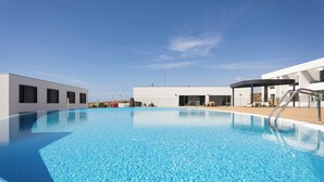 A modern building with a swimming pool, a covered seating area, and wind turbines in the background. - Home2Book Orballo Sea Views Apt, Terrace&Pool (Abades)