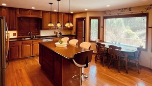 A modern kitchen with wooden cabinets, a central island with bar stools, pendant lighting, and a dining area with a large window. - Beautiful Four-Level Split Home @ Calm Cul-de-sac! (Roseville)