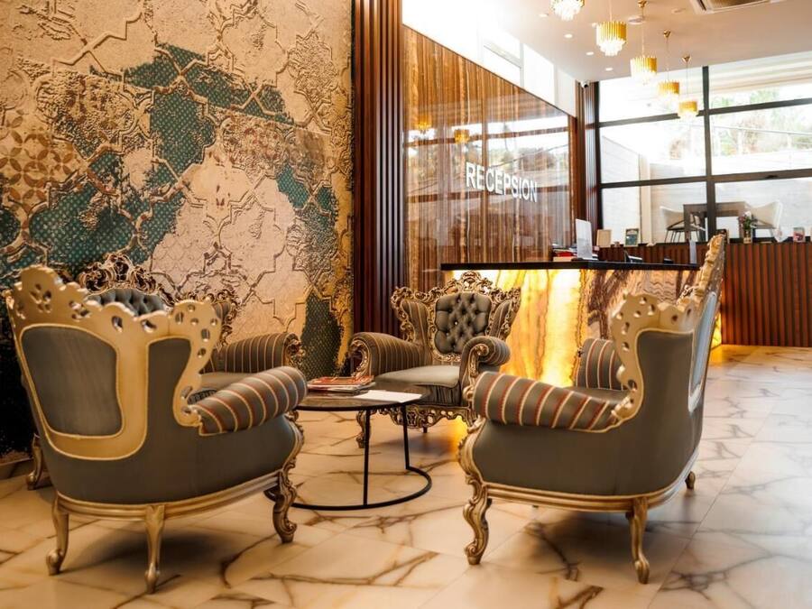 Uma área de recepção com piso de mármore, duas poltronas ornamentadas, um sofá estofado e um balcão de atendimento.