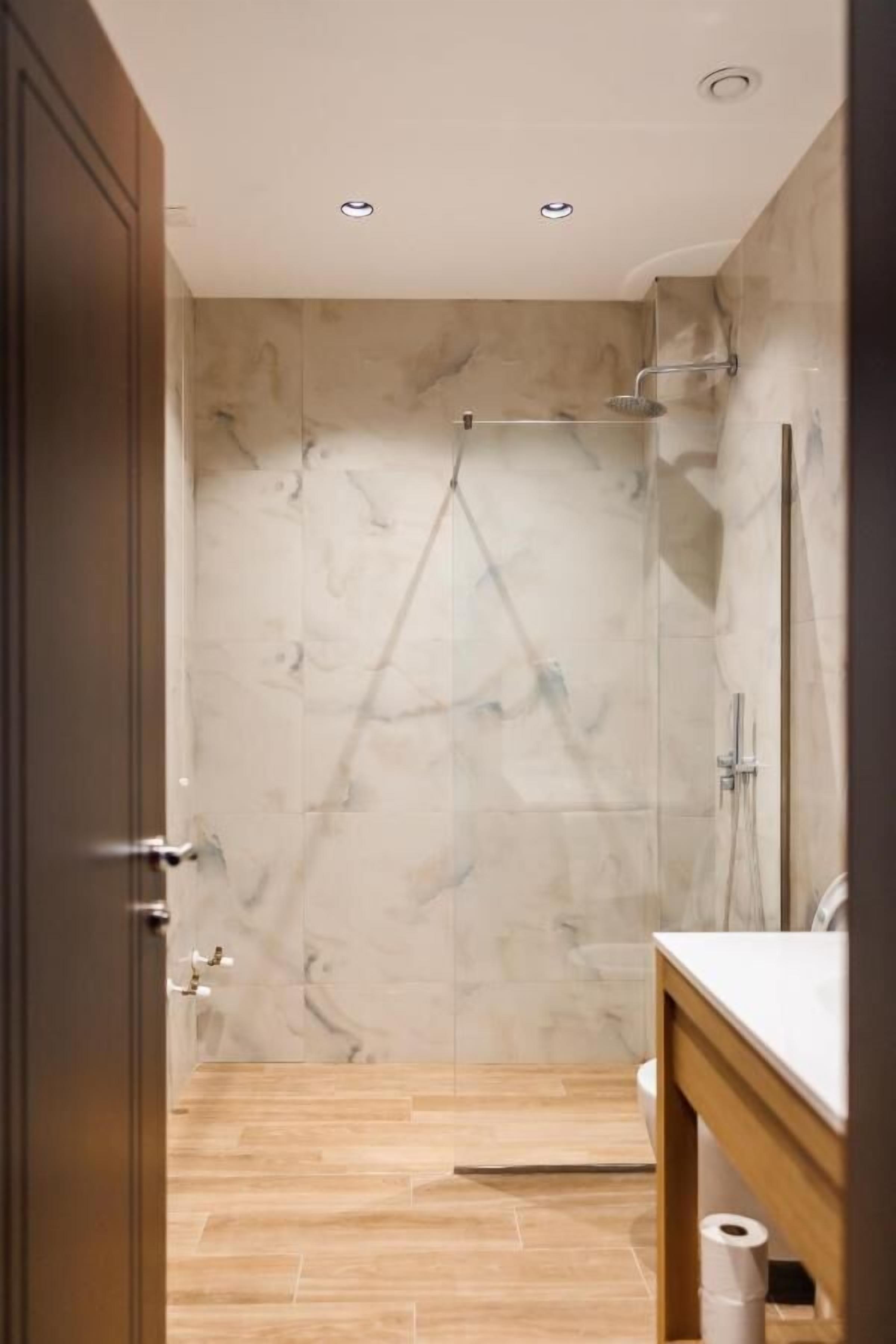 A modern bathroom with a shower, marble walls, a wooden floor, and a wooden door.