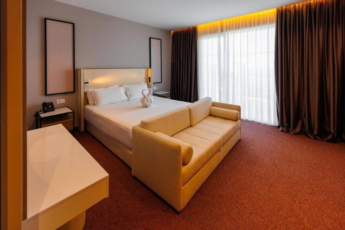 Una habitación de hotel con cama, sofá, escritorio y un ventanal con cortinas.