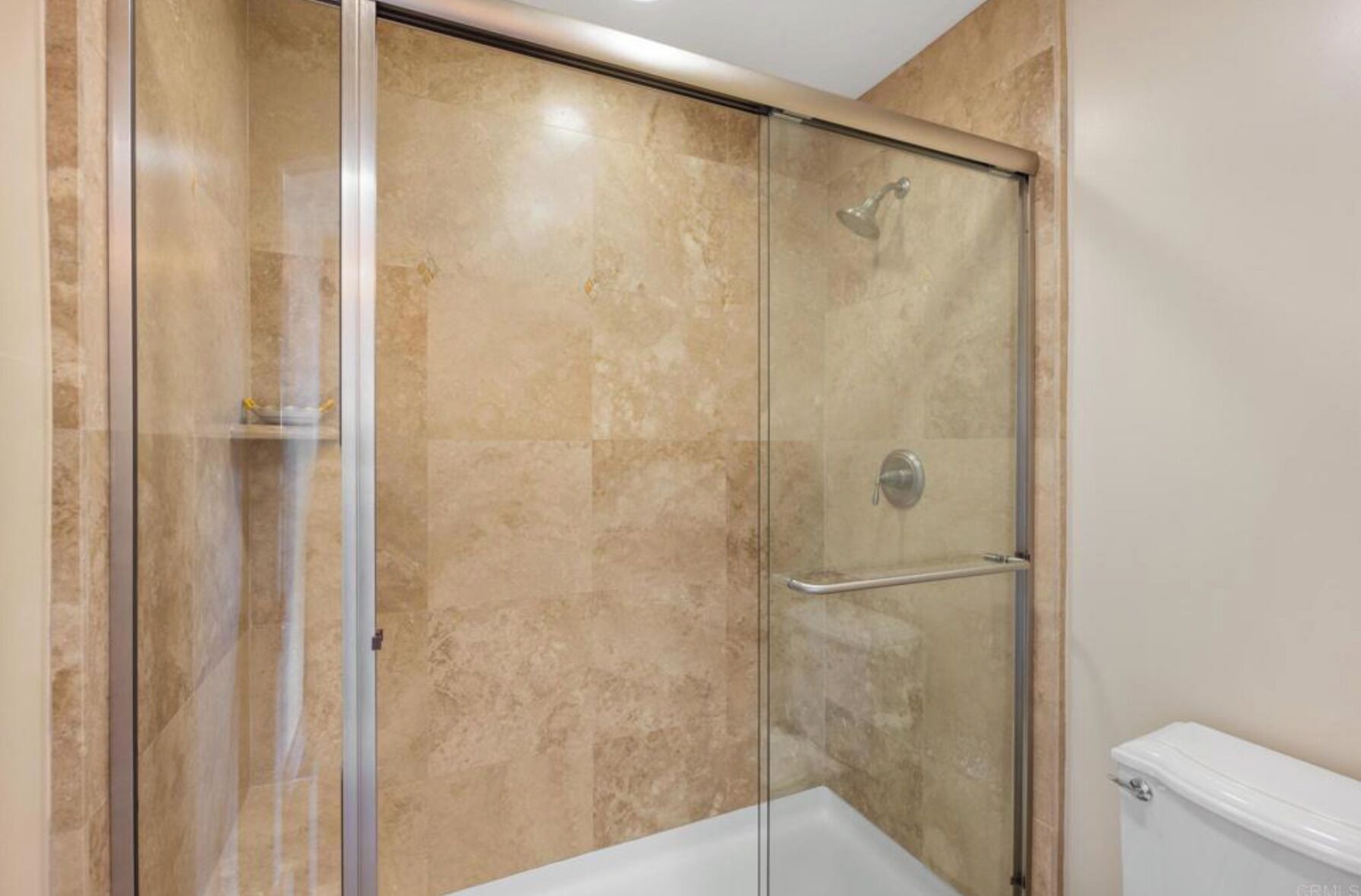 A glass-enclosed shower with a white bathtub, beige tiled walls, and a visible toilet.