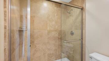 A glass-enclosed shower with a white bathtub, beige tiled walls, and a visible toilet.