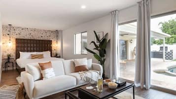 A modern hotel room with a bed, sofa, coffee table, and a view of the outdoor pool area.