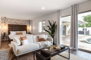 A modern hotel room with a bed, sofa, coffee table, and a view of the outdoor pool area.