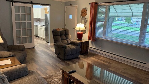 A living room with a sofa, armchair, and a coffee table. There is a sliding glass door leading to a kitchen area. A ceiling fan is visible, and a window with blinds is on the right side.