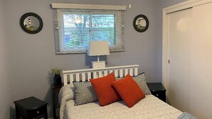 A bedroom with a bed, two bedside tables, a nightstand, a window with blinds, and two wall-mounted mirrors.