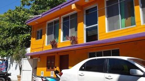 A two-story building with a yellow facade and purple roof, featuring multiple windows and a parked white car in front. - Private room for 5 people. A/C, TV, Wi-Fi, minibar, kitchen and living. Downtown | Isla Mujeres  #2 (Isla Mujeres)