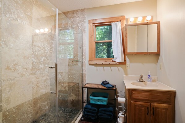 Baño con cabina de ducha de vidrio, tocador de madera y ventana con cortina.