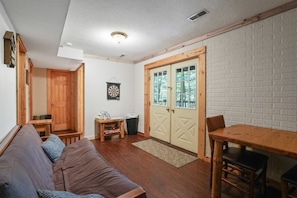 A cozy living room with a wooden sofa, a dining table with chairs, a dartboard, and a door leading to a balcony.