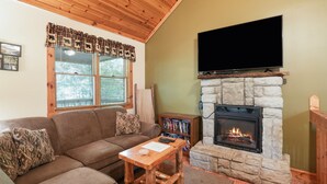 Una sala de estar acogedora con chimenea de piedra, un televisor de pantalla plana montado encima y techo de madera, además de una ventana con persianas.