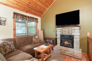 A cozy living room with a stone fireplace, a flat-screen TV mounted above it, a wooden ceiling, and a window with blinds.
