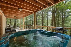 Un área de jacuzzi con cubierta de madera, rodeada de árboles, con mobiliario exterior y una parrilla.