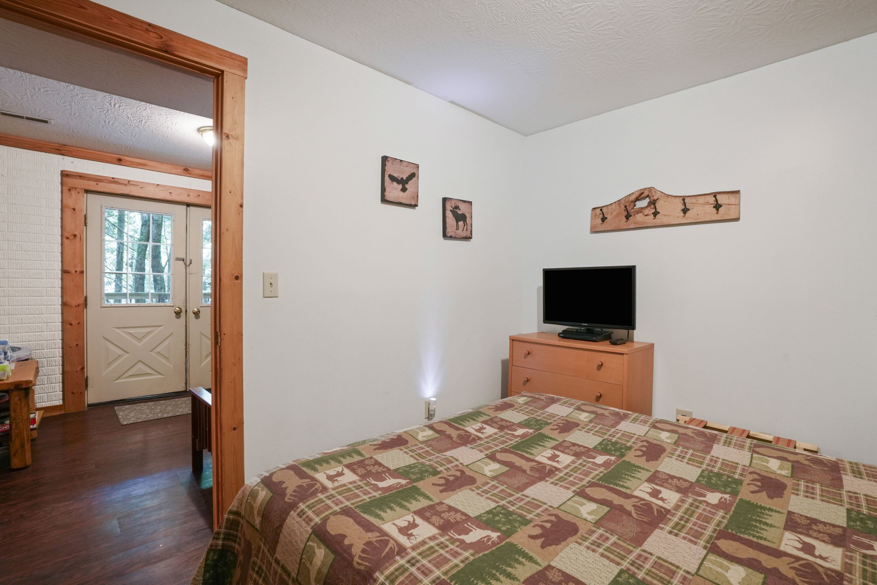 Ein Schlafzimmer mit einem Bett, einem Fernseher auf einer Holzkommode, einer Tür zu einem anderen Raum und zwei an der Wand befestigten Bildern.