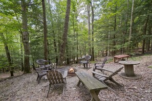 Un camping en el bosque con una fogata, bancos de madera y sillas de plástico.