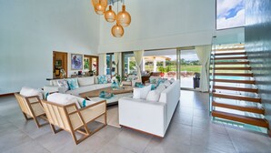 A spacious living room with a large sofa, a coffee table, and wicker chairs. There are hanging wicker lamps, a large window with a view of a pool, and a sliding glass door leading to an outdoor patio.