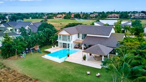 A large two-story house with a swimming pool, surrounded by lush greenery and other houses.