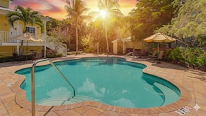 A swimming pool with a sunlit sky, palm trees, and a building with a balcony. - Gertrude's Siesta | Pool, Jacuzzi! 1-minute to Beach! 4-min Dining & Music! (Siesta Key)
