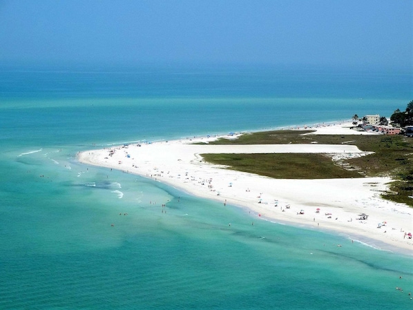 A coastal beach with a wide sandy area, clear turquoise waters, and a few buildings in the background.