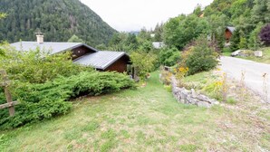 Eine rustikale Holzhütte mit geneigtem Dach, umgeben von üppiger Vegetation und einer Steinmauer.