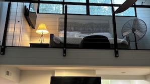 Camera da loft moderna con una grande finestra, un letto, una lampada da comodino e una ventola. Più in basso, è presente un angolo cottura con una TV, una lavatrice e un camino.