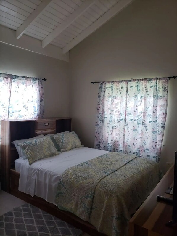 Une chambre à coucher comprenant un lit, une table de chevet, une commode et une fenêtre agrémentée de rideaux à motifs floraux.