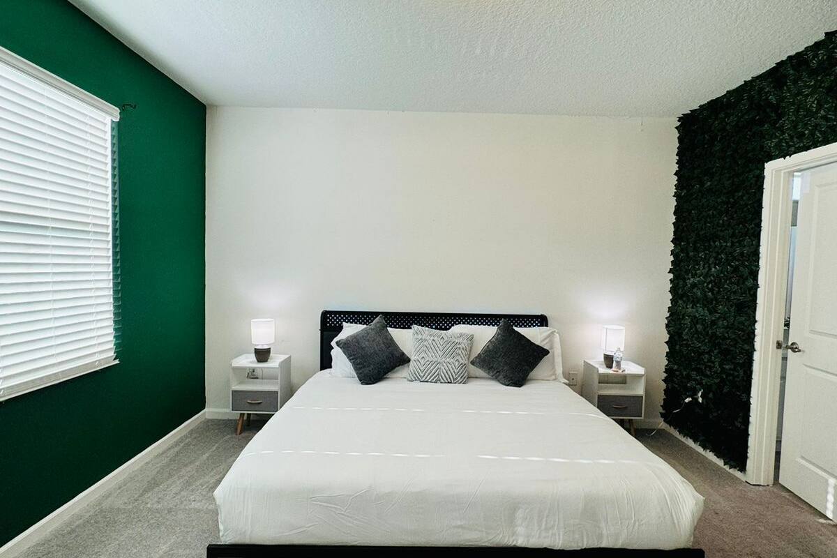 A bedroom with a bed, bedside tables, a wall-mounted light fixture, a window with blinds, and a green wall.