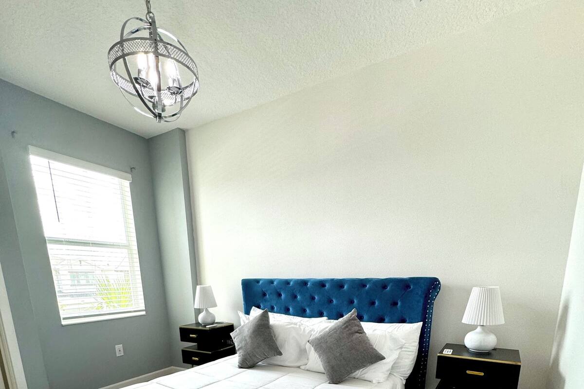 A bedroom with a large bed, two bedside tables, a hanging light fixture, and a window with blinds.