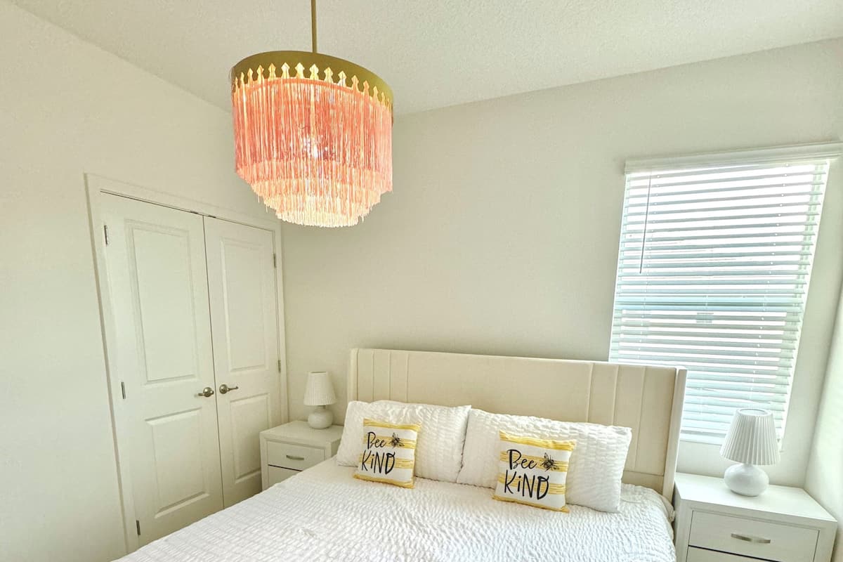 A bedroom with a bed, a nightstand, a dresser, a chandelier, and a window with blinds.
