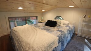 Un dormitorio con techo de madera, una cama con colcha estampada, una ventana con cortinas y una mesita de noche con lámpara.