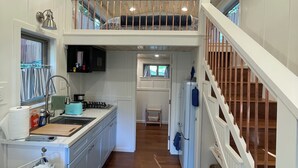 Una cocina amplia con una isla central, un comedor con una silla azul y una escalera que lleva a un dormitorio en el altillo.