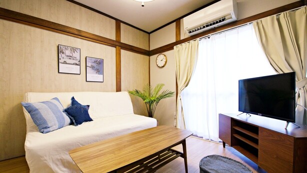 A room with a white sofa, a wooden coffee table, a TV on a wooden stand, a plant, and a clock on the wall. - -Takaya house- Minamirokugou close Haneda AP (Ota City)