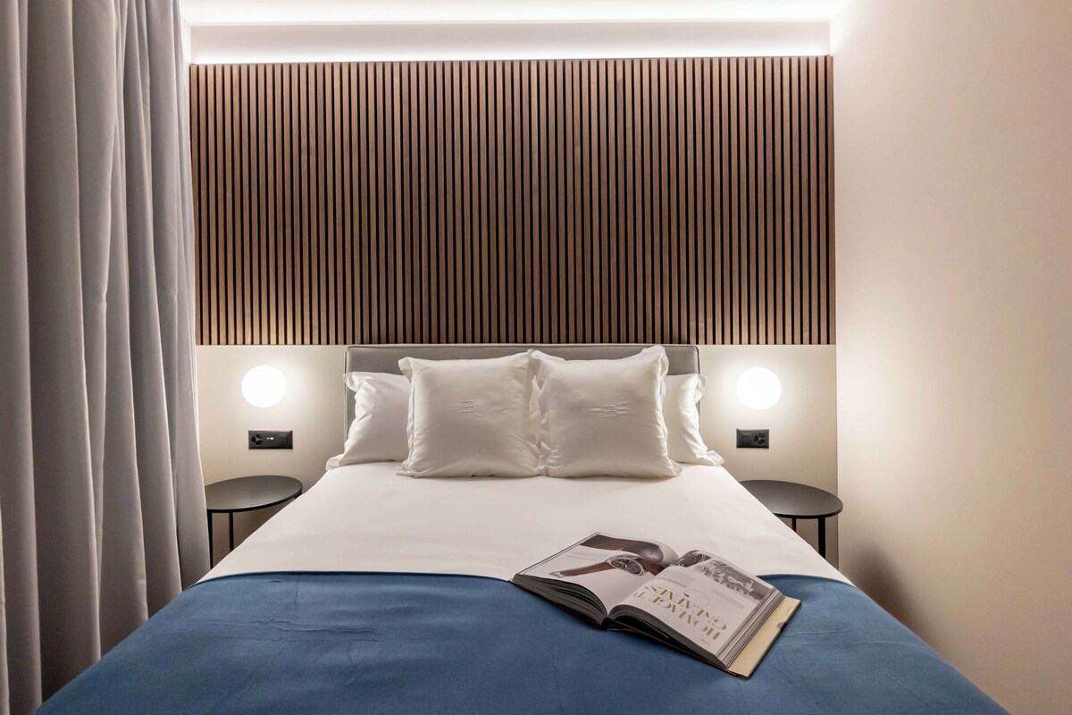 Quarto de hotel com cama, duas mesas de cabeceira, luminária na parede e cortina.