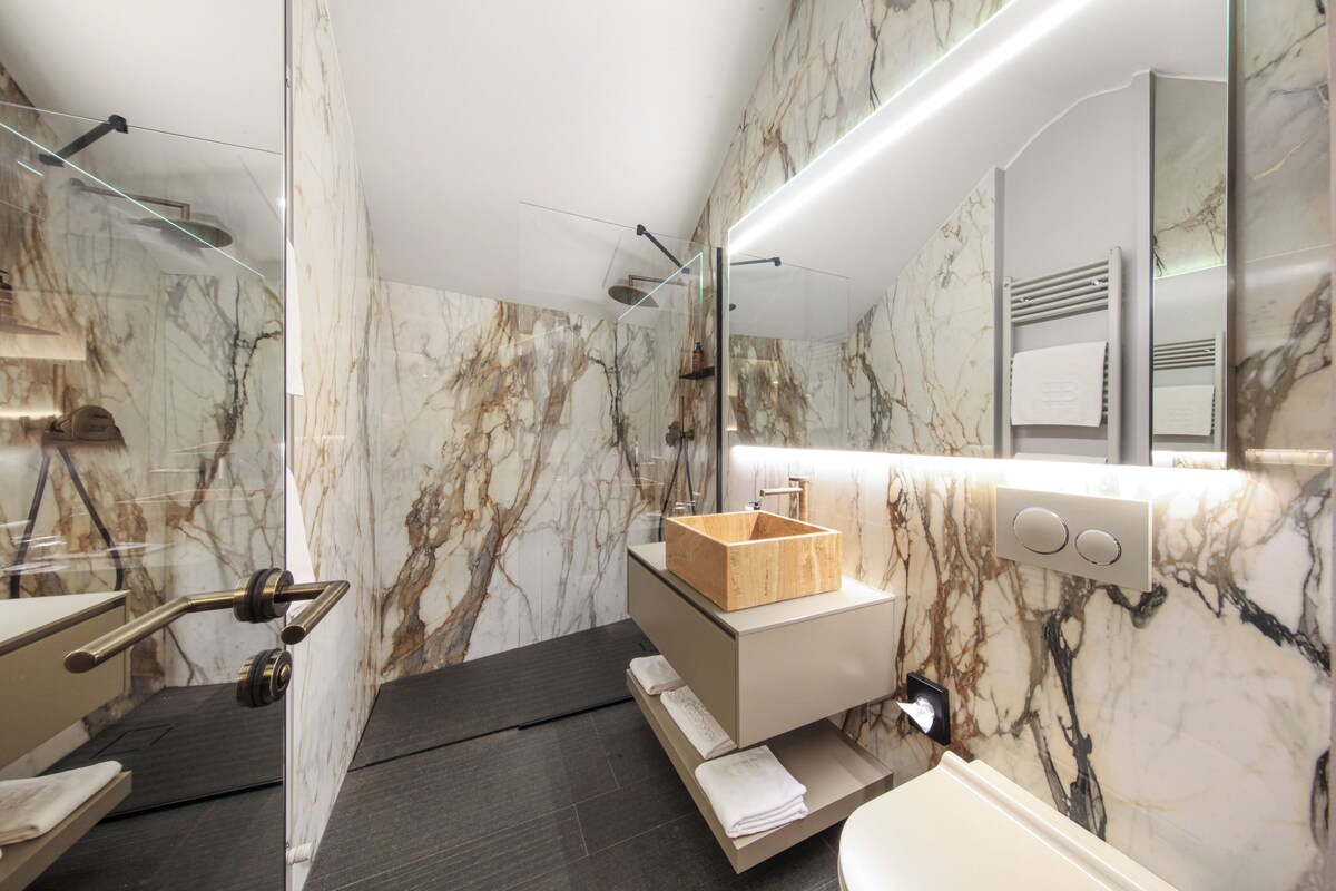 A modern bathroom with a marble wall, a wooden vanity, a glass shower enclosure, and a built-in towel rack.