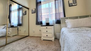 Une chambre à coucher avec un lit, une table de chevet, un miroir, une commode et une fenêtre avec des rideaux.