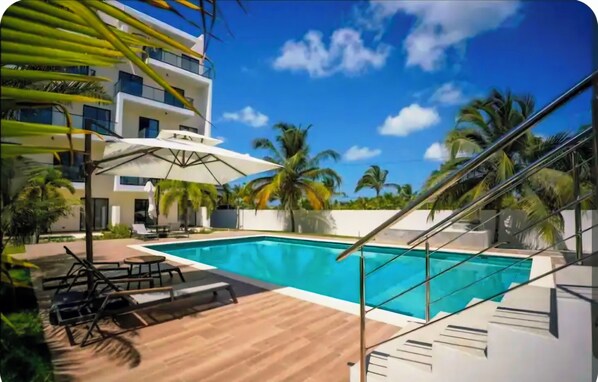 A modern hotel with a swimming pool, palm trees, and a clear blue sky.