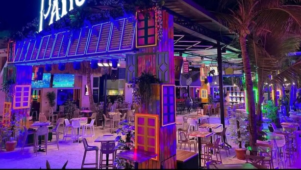 Ein Außenbereich mit Essbereich, Tischen, Stühlen und einem Neon-Schild mit der Aufschrift "Pal Patio". Außerdem gibt es einen überdachten Sitzbereich mit einem Reetdach.