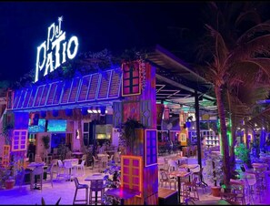 An outdoor dining area with tables and chairs, a neon sign reading "Pal Patio," and a covered seating area with a thatched roof.