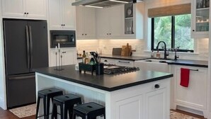 A modern kitchen with white cabinetry, a central island with stools, a stainless steel range hood, and a window providing natural light.