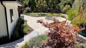 A modern house with a stone patio, a fire pit, and a garden with various plants and trees.