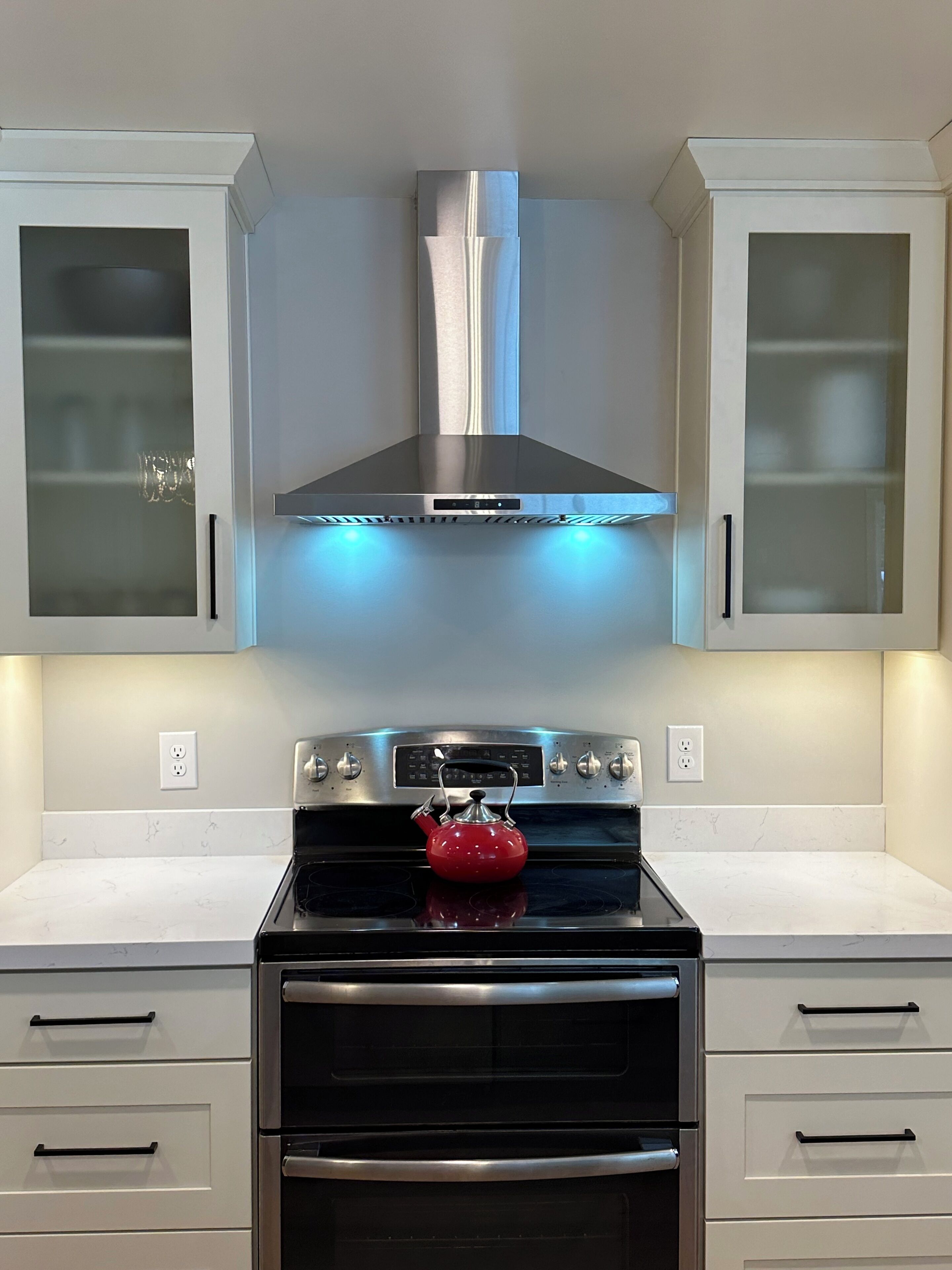 A kitchen with a stainless steel range hood, a red kettle on the stove, and white cabinets with glass doors.