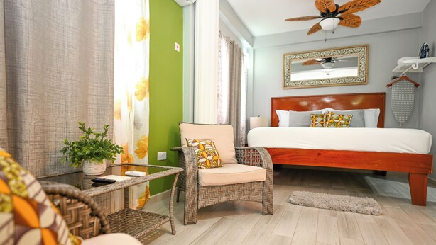 Ein Schlafzimmer mit Bett, Stuhl, kleinem Tisch mit Pflanze, Deckenventilator und Spiegel an der Wand.