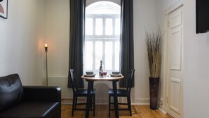 Une pièce avec un canapé en cuir noir, une table ronde en bois accompagnée de deux chaises, une plante en pot et une grande fenêtre avec des rideaux.