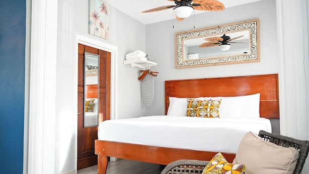 A bedroom with a wooden bed, a ceiling fan, a mirror, and a wicker chair. - Ocean Villa (Corinth)