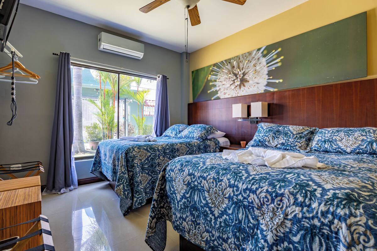 A hotel room with two beds, a ceiling fan, an air conditioning unit, a large window with curtains, and a view of greenery outside.