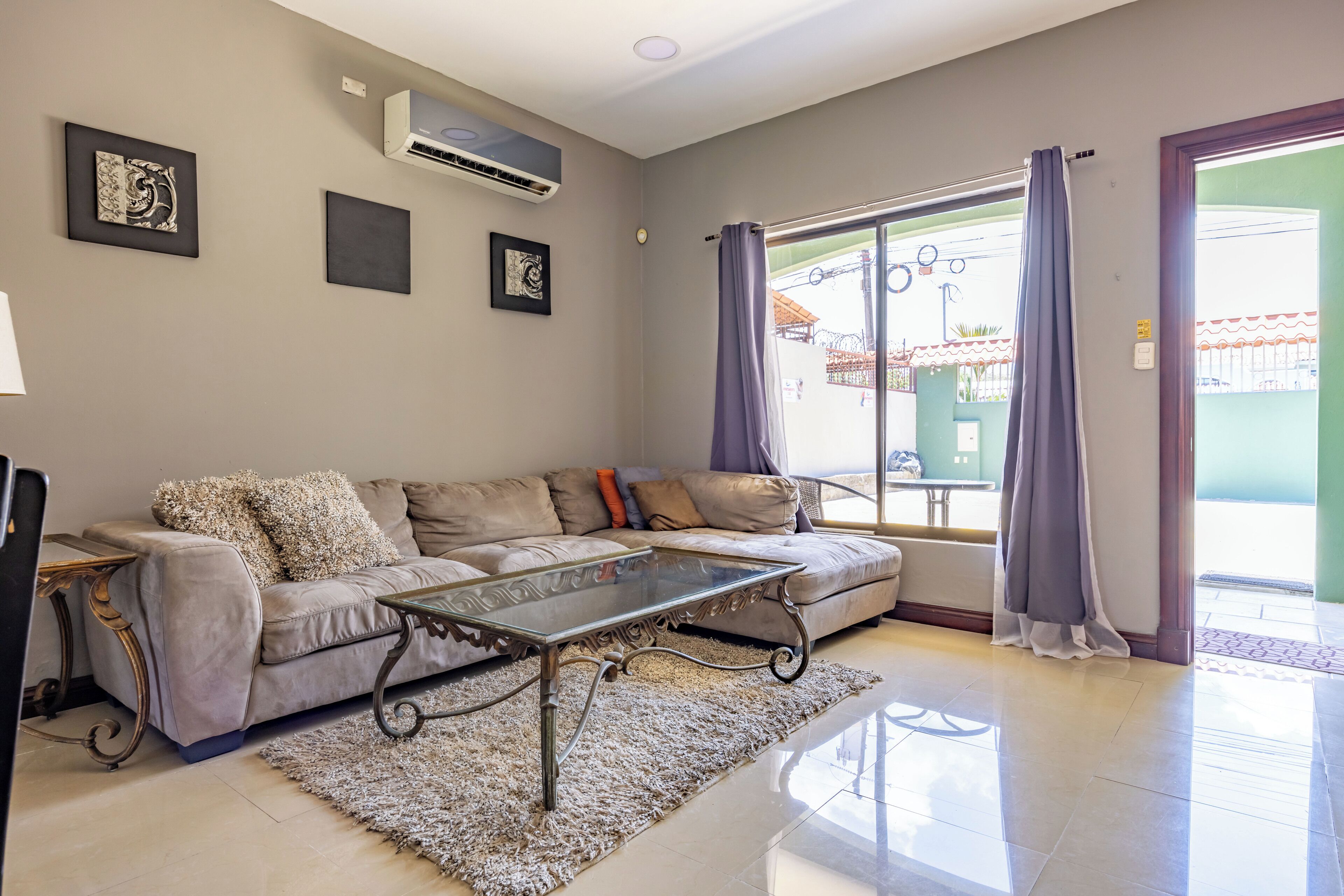 A living room with a sofa, a glass coffee table, and a large window with curtains.